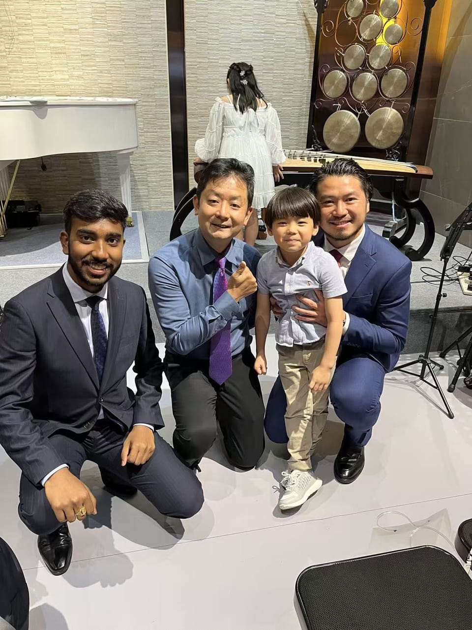Three men in suits kneel in front of a small stage with a young boy standing between two of them, while a girl in a white dress, seen from behind, stands at a guzheng.