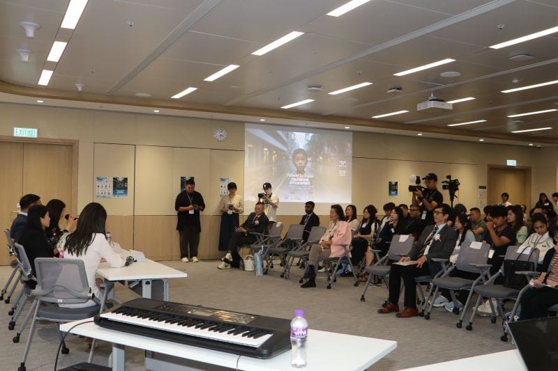 An audience on rows of chairs listens to speakers seated at a table on the left, with an electronic keyboard in the foreground and a camera crew filming from the back of the room.