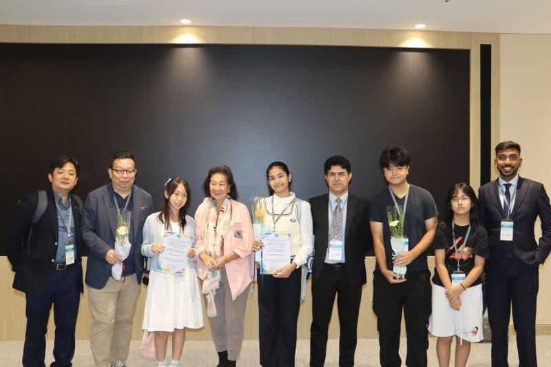 Nine people stand in a row in front of a dark wall, some of the younger ones holding certificates and single flowers.