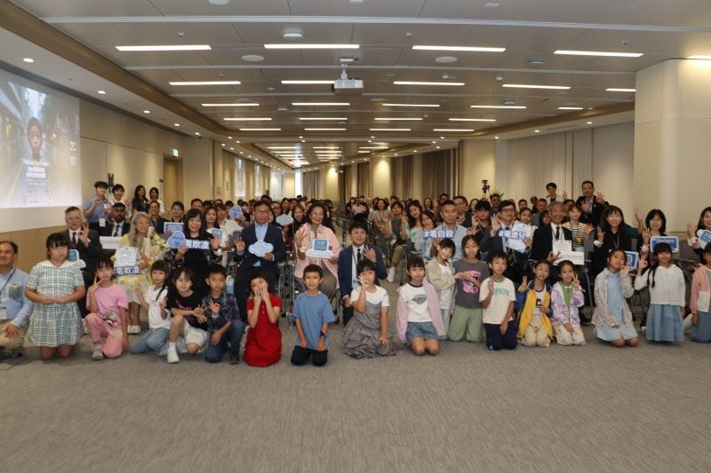A full conference room poses for a group photograph, with children sitting on the floor at the front and adults behind them, some holding small signs.