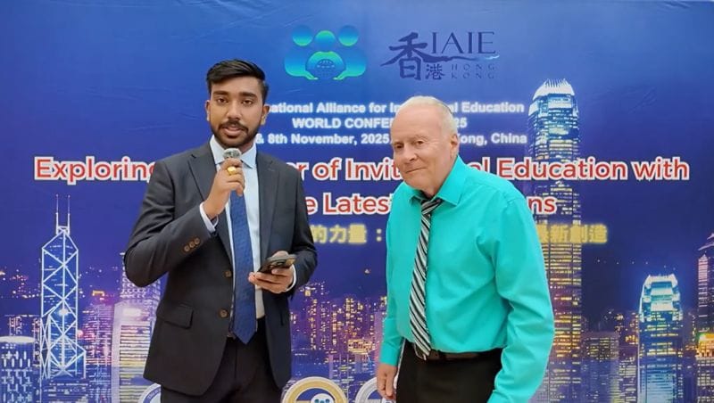 Zwei Männer stehen vor einer Konferenzrückwand mit der Skyline von Hongkong: Der eine, im Anzug, hält ein Mikrofon, der andere trägt ein türkisfarbenes Hemd und eine gestreifte Krawatte.