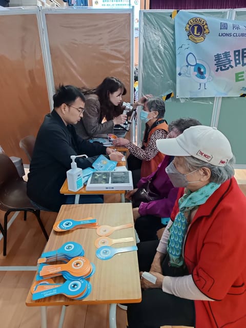 At a row of school desks, a woman examines a seated visitor’s eye with a hand-held instrument while a man beside her writes, and more visitors wait in the foreground, the nearest wearing a cap and a face mask.