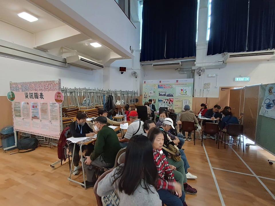 A school hall with visitors seated in rows, volunteers at desks filling in forms with them, and display boards along the far wall.