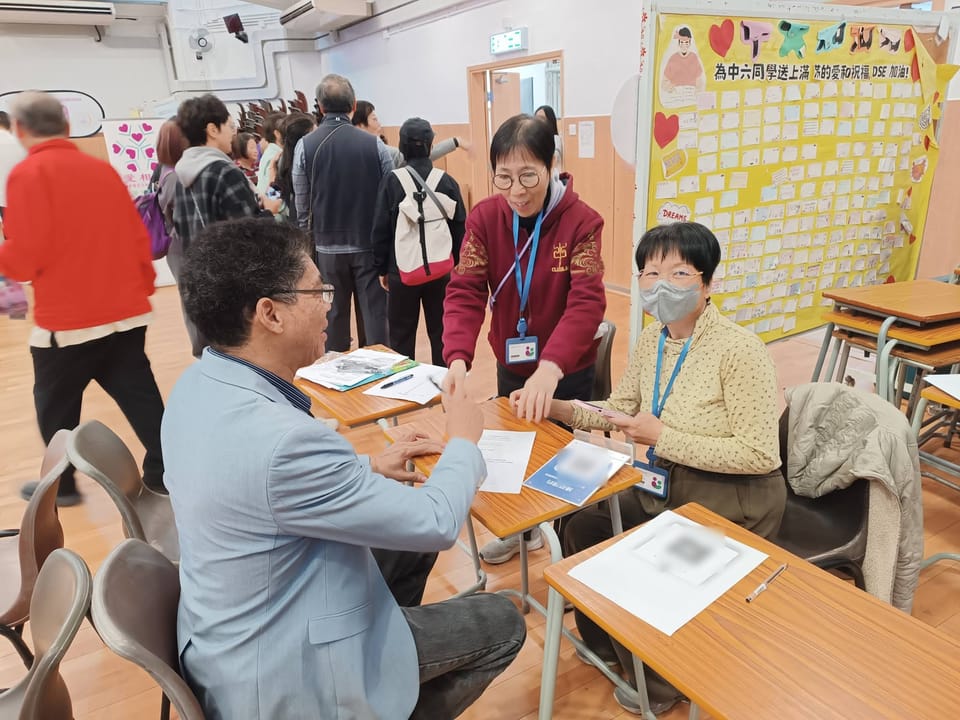 A volunteer wearing a lanyard leans across a school desk, smiling, towards a seated visitor, while a second volunteer in a face mask sits beside her holding a phone.