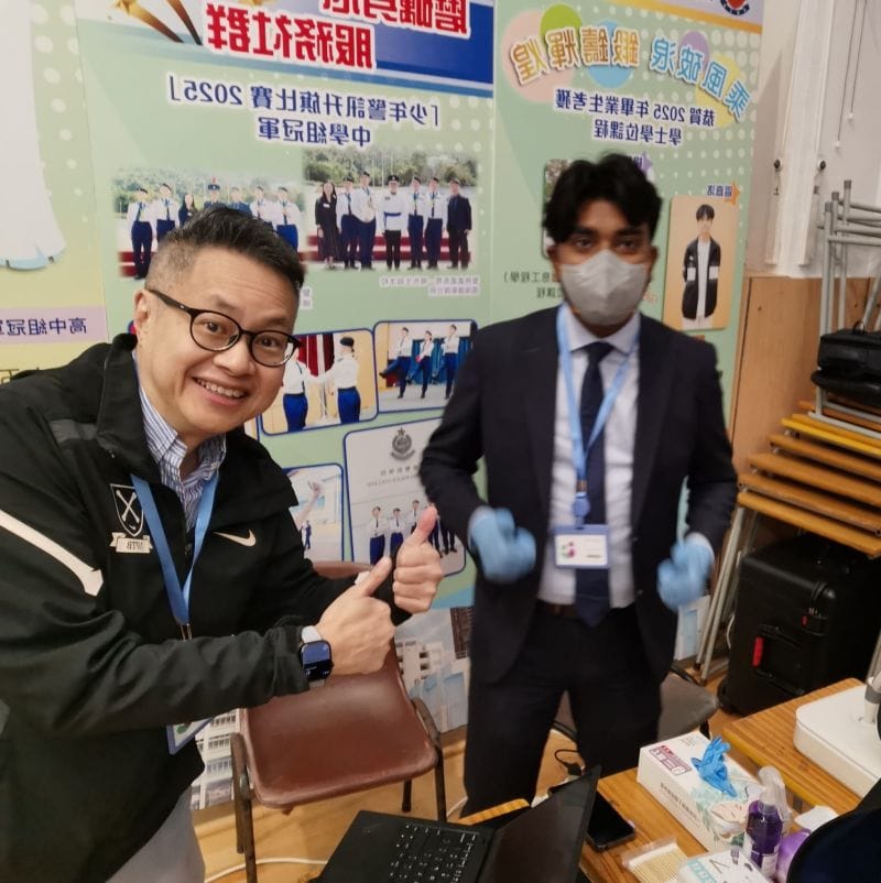 Two volunteers at a screening station: one, seated, grins and gives two thumbs up, and the other stands behind him in a suit, a face mask and blue gloves.