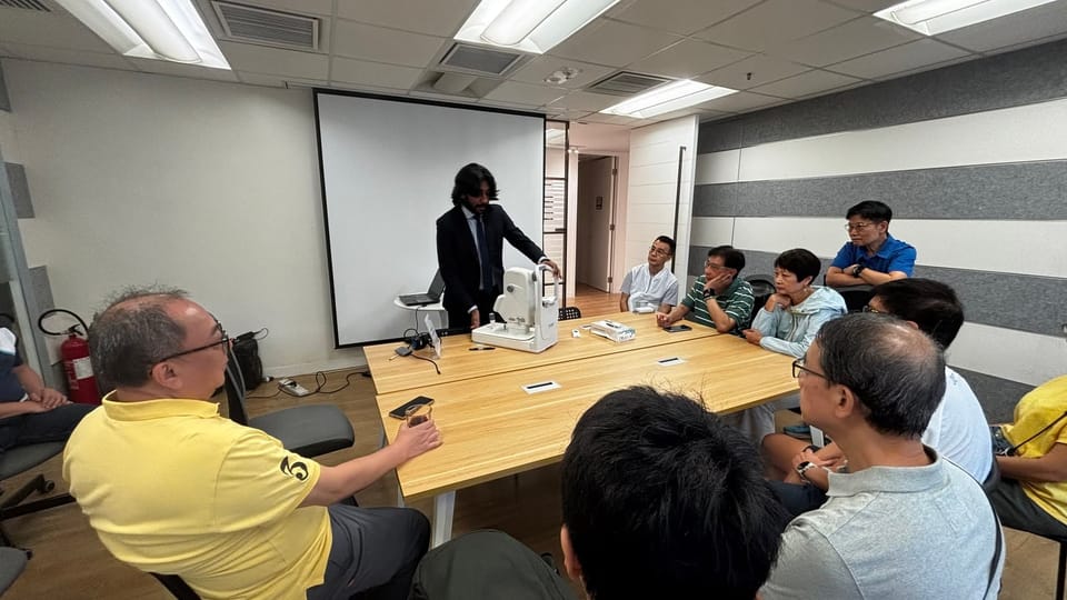 A man in a suit stands at the head of a long table beside a fundus camera, speaking to people seated around it.
