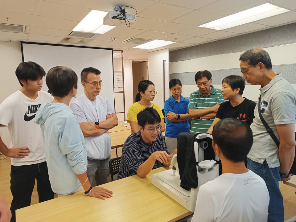 A seated trainer in a striped top explains a fundus camera to a ring of people standing round the table, as one of them sits facing the camera.