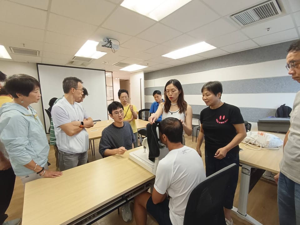A trainer lifts the dark cloth of a fundus camera for a man seated at it, while a seated colleague explains and others stand watching in a meeting room.