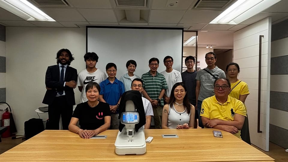 Thirteen people pose in two rows behind a table with a fundus camera on it, in front of a projection screen.