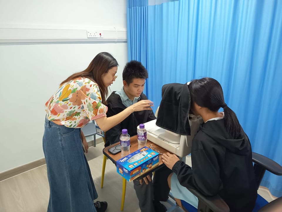 A trainer leans in to guide a student in school uniform at the controls of a fundus camera while a second student looks into it under a dark cloth.