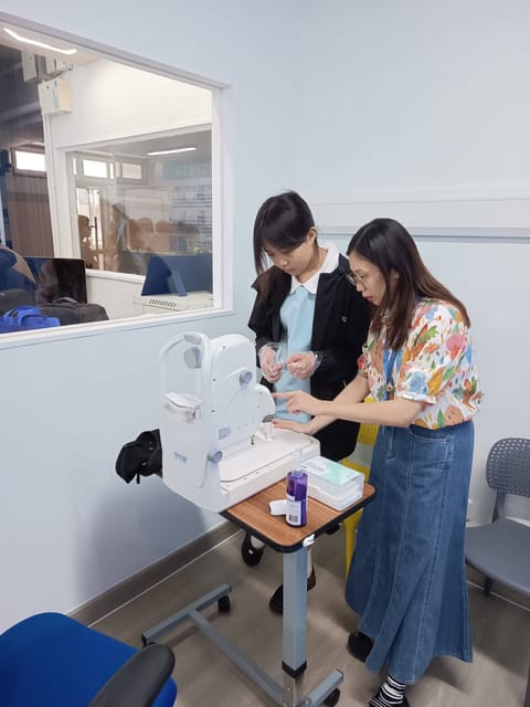 A trainer points to the base of a fundus camera on a trolley while a student in school uniform and disposable gloves stands beside her, looking down at it.