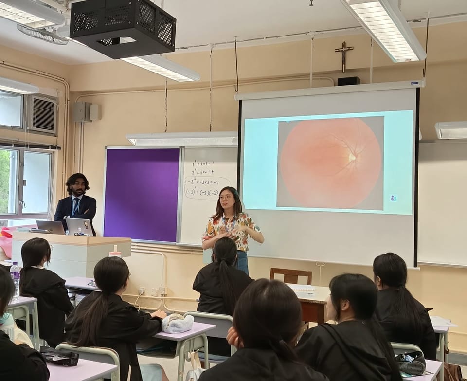 Students at their desks, seen from behind, face a trainer who is speaking beside a projected photograph of a retina, while a man in a suit stands at the teacher’s desk.