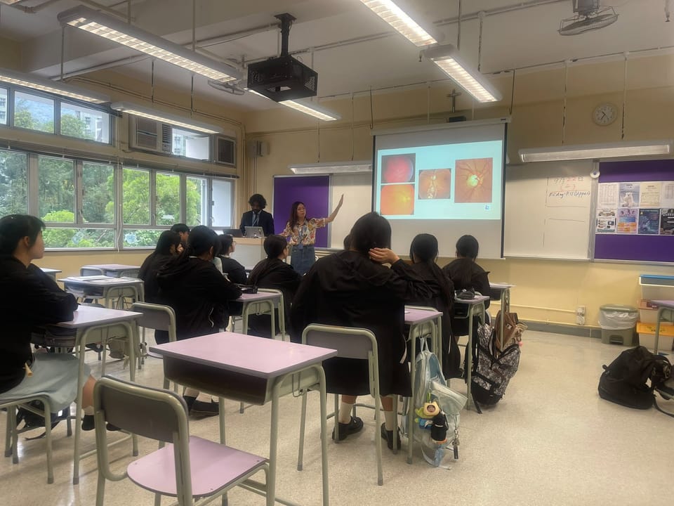 A classroom seen from the back: students at their desks watch a trainer who raises her arm towards a screen showing photographs of retinas.