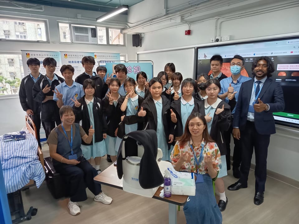 A class of students in school uniform and four adults pose around a fundus camera on a table, many giving a thumbs-up or a V sign.