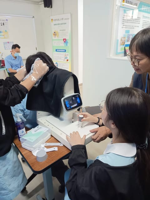 A student in school uniform works the joystick of a fundus camera with an adult beside her, while a classmate sits at the chin rest under a dark cloth that gloved hands hold in place.