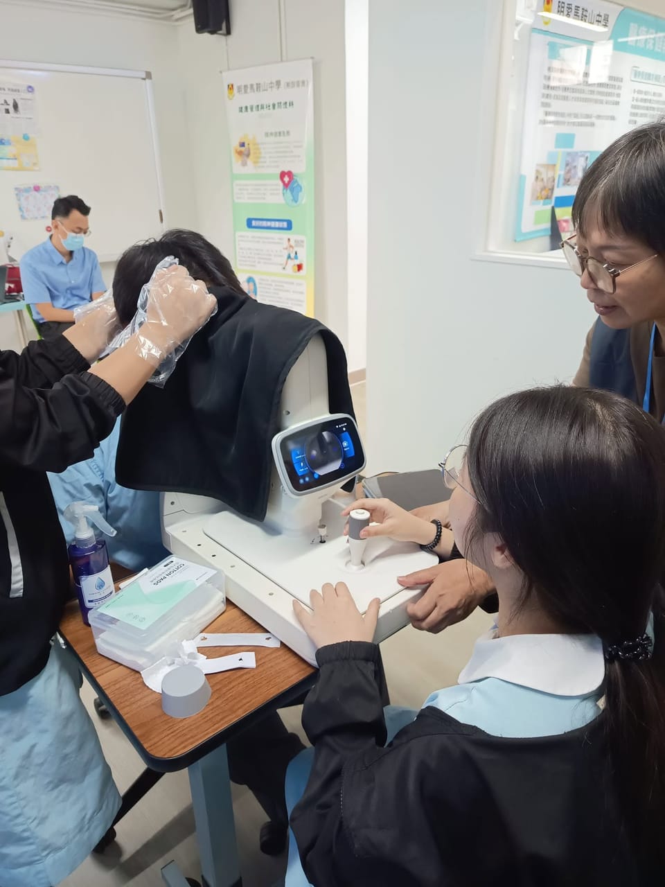 A student in school uniform works the joystick of a fundus camera with an adult beside her, while a classmate sits at the chin rest under a dark cloth that gloved hands hold in place.